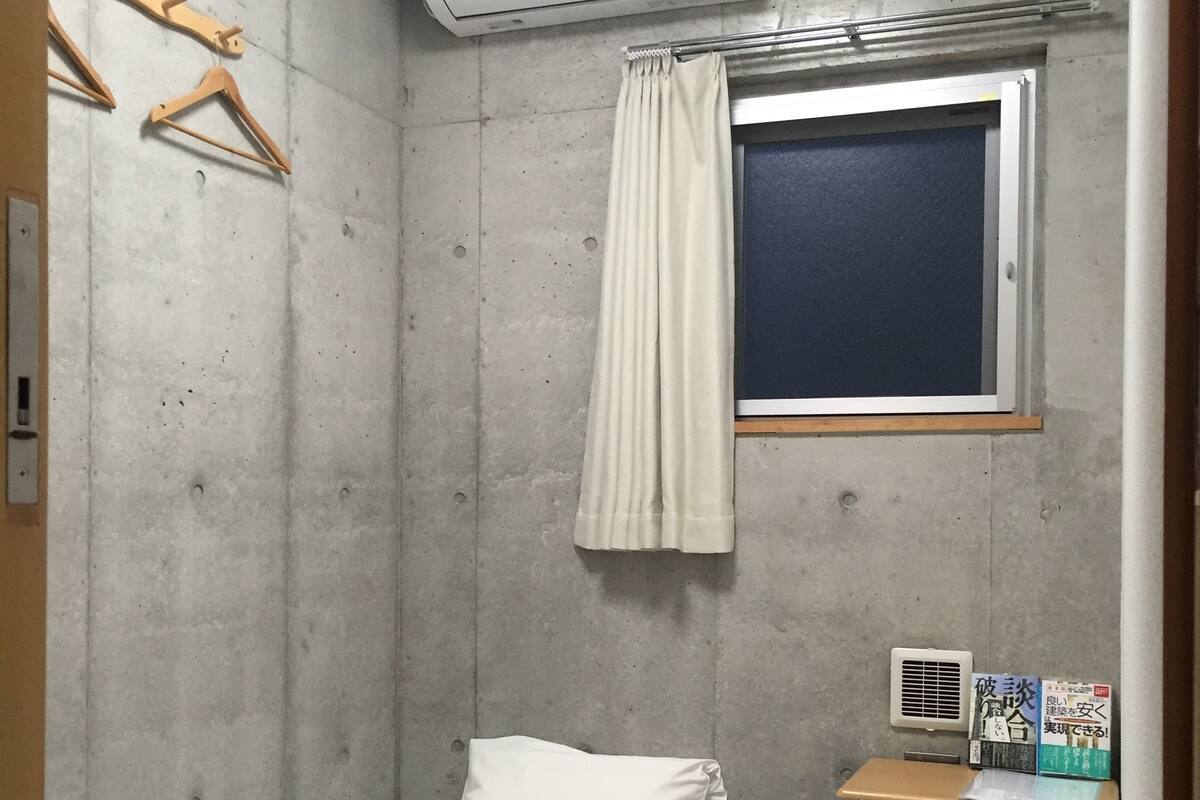 A small, compact room with a single bed, a window with curtains, an air conditioner, a small table with a trash bin, and a wall-mounted heater.