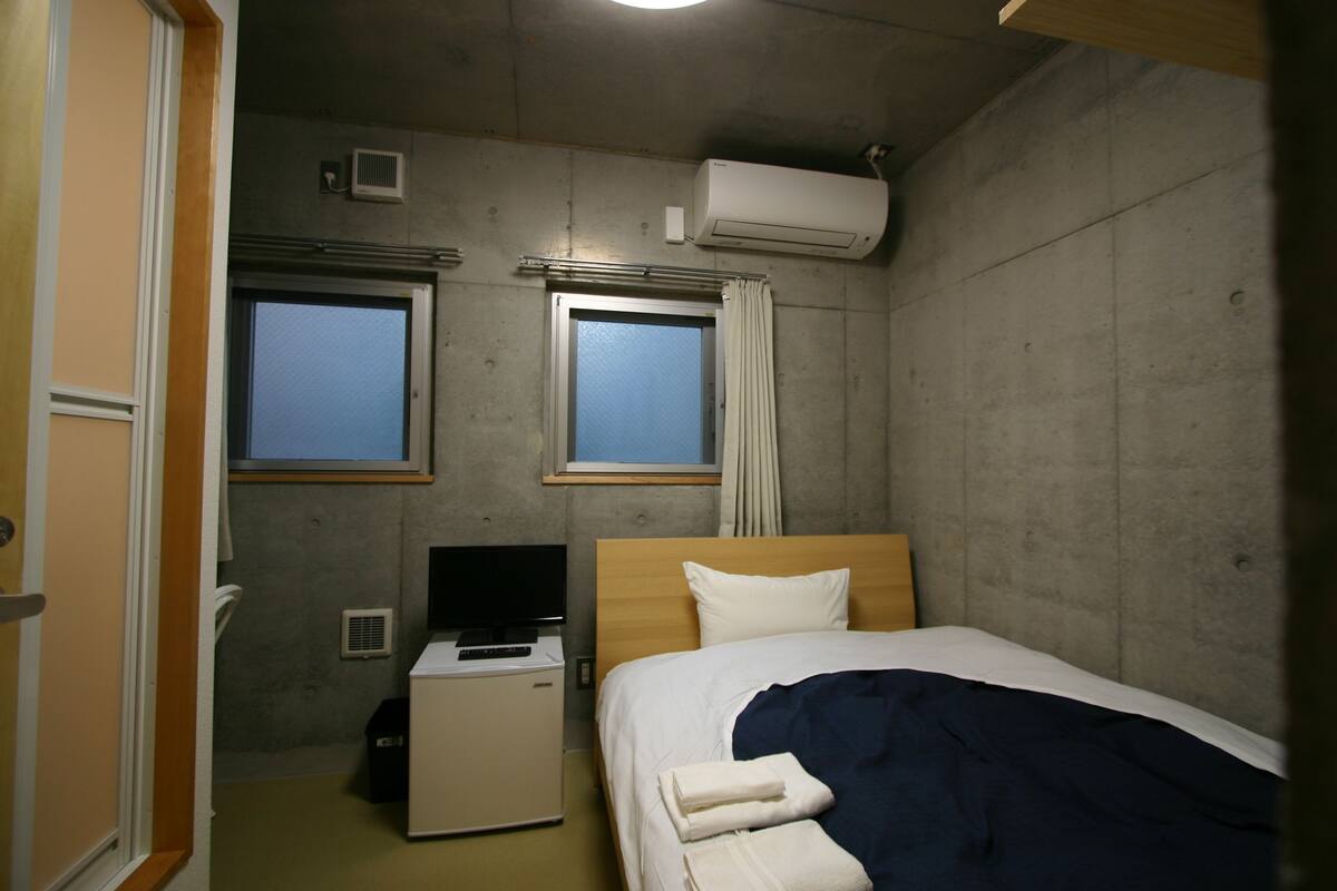 A compact hotel room with a single bed, a small desk with a computer, and two windows.