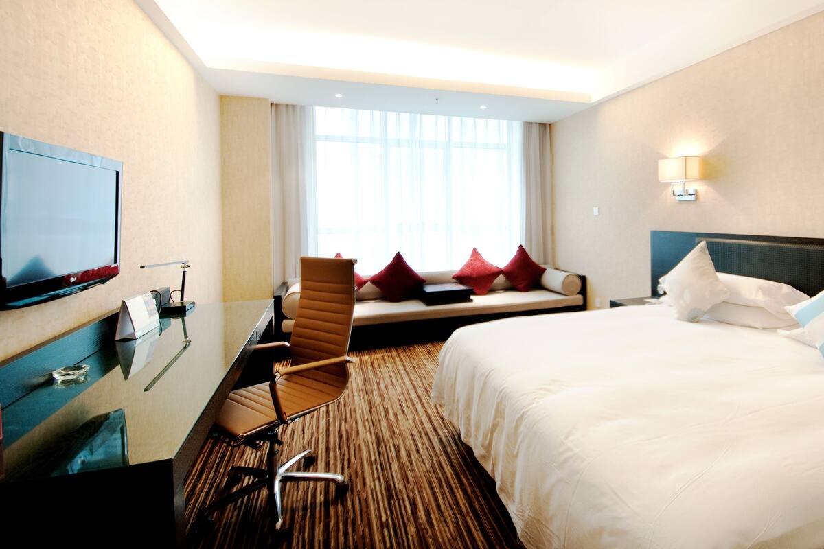 Habitación de hotel con cama, escritorio, silla, televisor y ventana con cortinas.