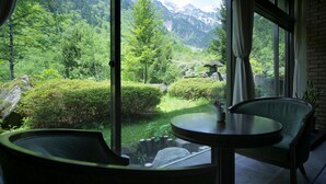 Een kamer met een groot raam dat uitzicht biedt op een weelderig groen landschap met bergen op de achtergrond. Op de voorgrond staan een ronde tafel en twee stoelen.