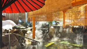 Een traditionele Japanse onsen met houten constructie, een rode paraplu en een stenen bad omringd door rotsen.
