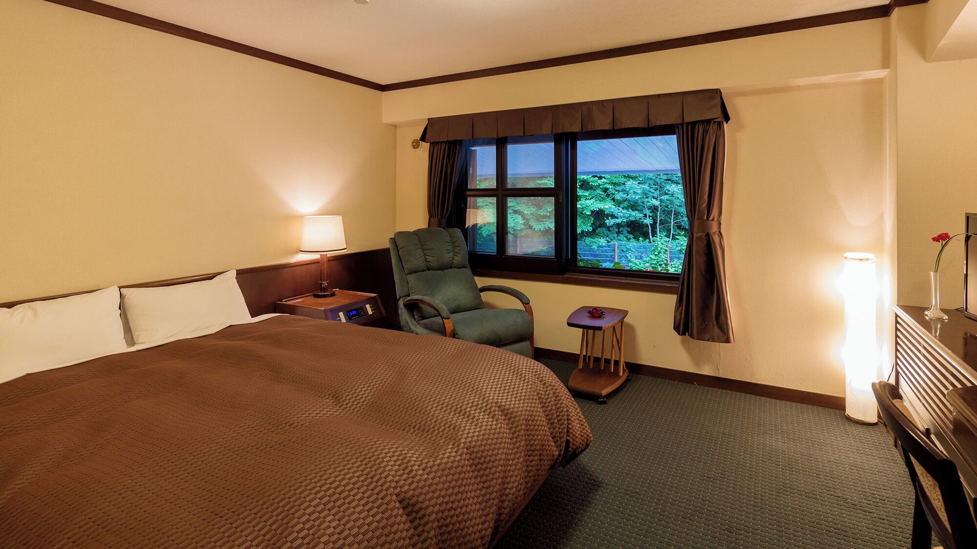 A hotel room with a large bed, a chair, a small table, a nightstand, a window with curtains, and a lamp.