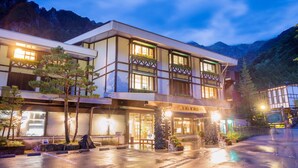 Een hotel in traditionele bouwstijl, met meerdere balkons met planten, een goed verlichte entree en uitzicht op bergen.