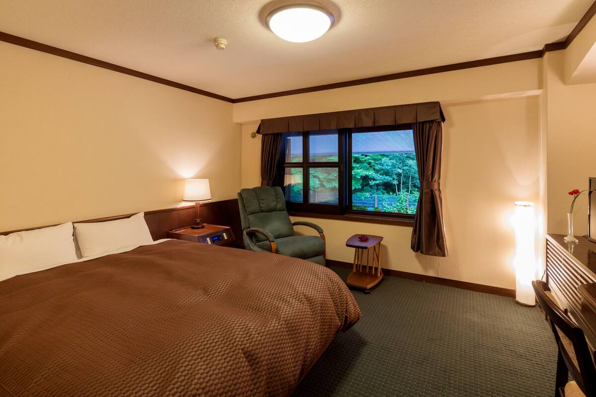 Habitación de hotel con una cama grande, una silla, una mesita, una lámpara y una ventana con vistas al verdor.