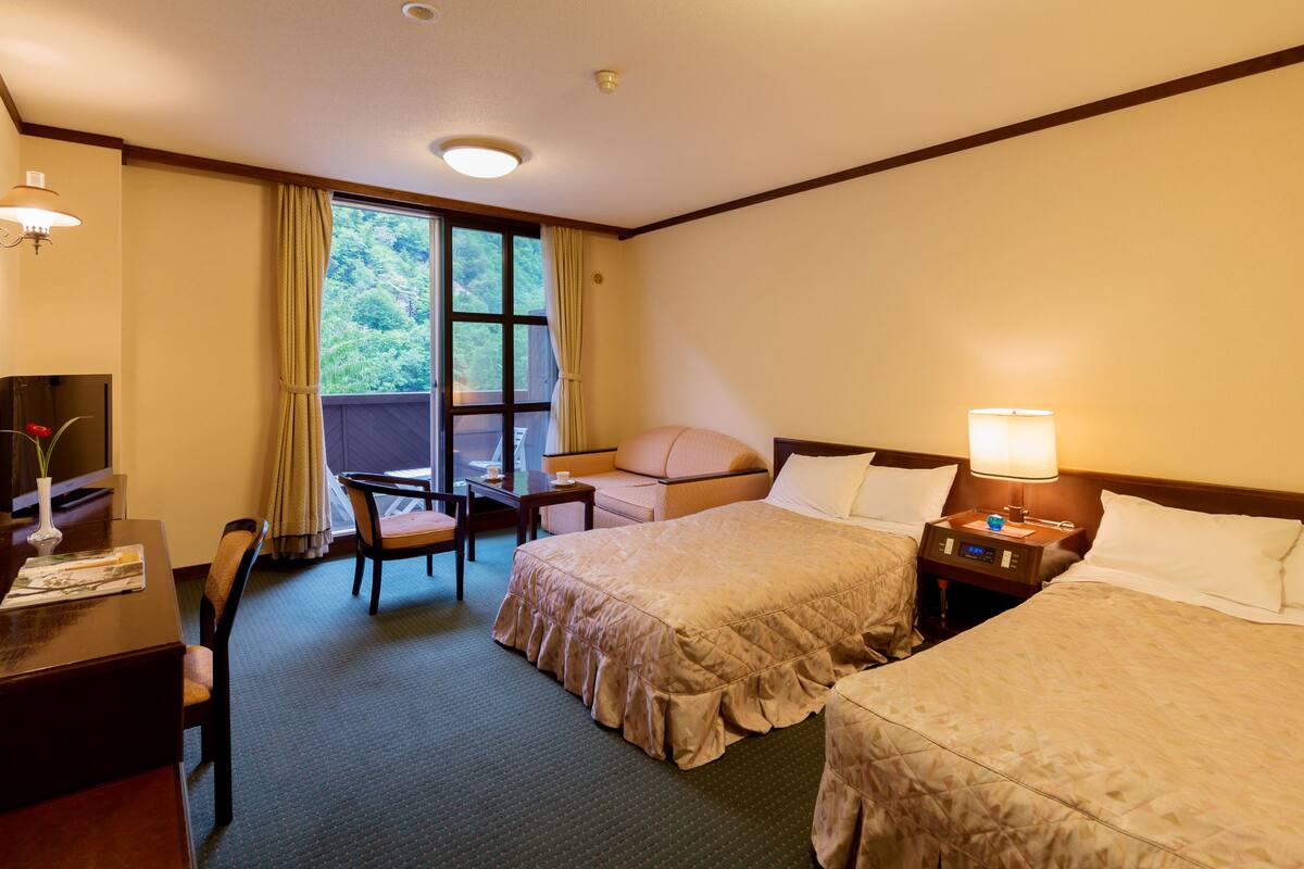 Habitación de hotel con dos camas, un escritorio con silla, una mesita y una ventana con cortinas.