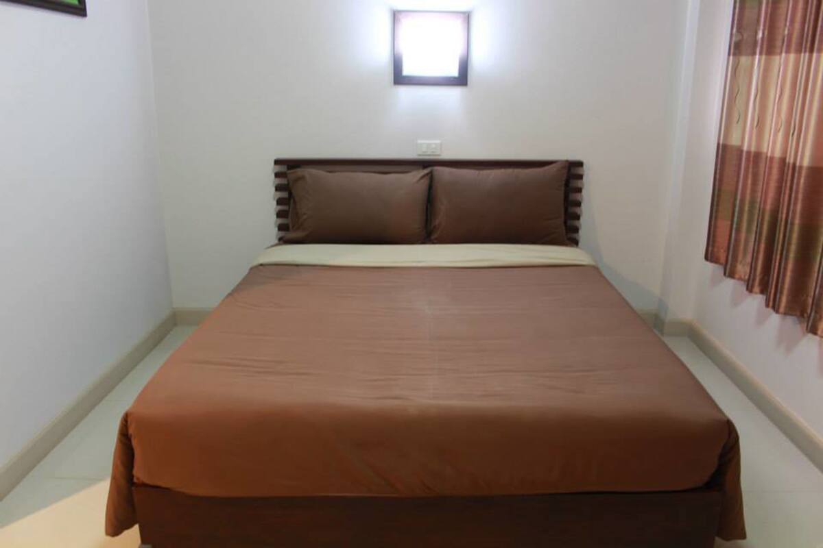 A single-bed room with a brown bedspread, a headboard, and a window with curtains.