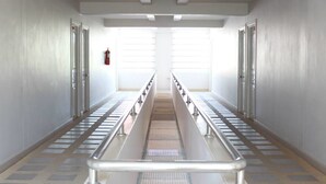 A long, narrow hallway with a central escalator flanked by metal railings, leading to a bright, open area. - Pornkasem House (Chiang Rai)