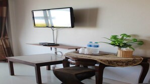 A room with a TV mounted on the wall, a wooden table with a bench, a small round table with a plant, and a trash bin. - Pornkasem House (Chiang Rai)
