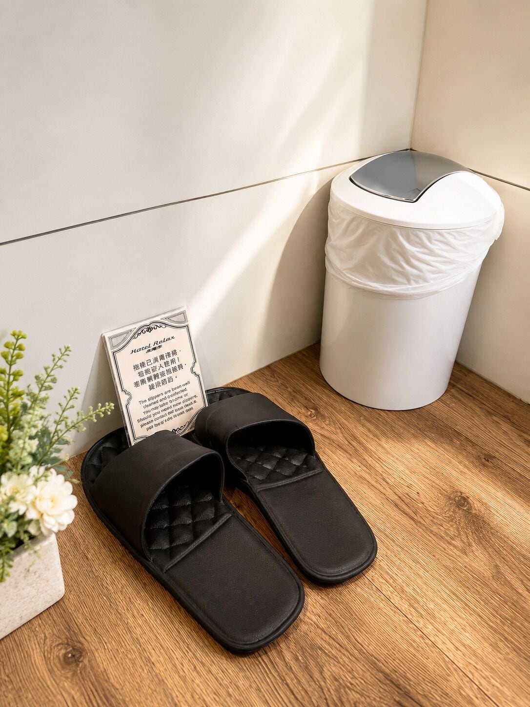 Sorte slippers på en træoverflade ved siden af en hvid skraldespand.