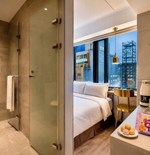 Habitación de hotel moderna con cabina de ducha de vidrio, cama con ropa blanca, mesita de noche con lámpara y vistas a la ciudad a través de amplias ventanas.