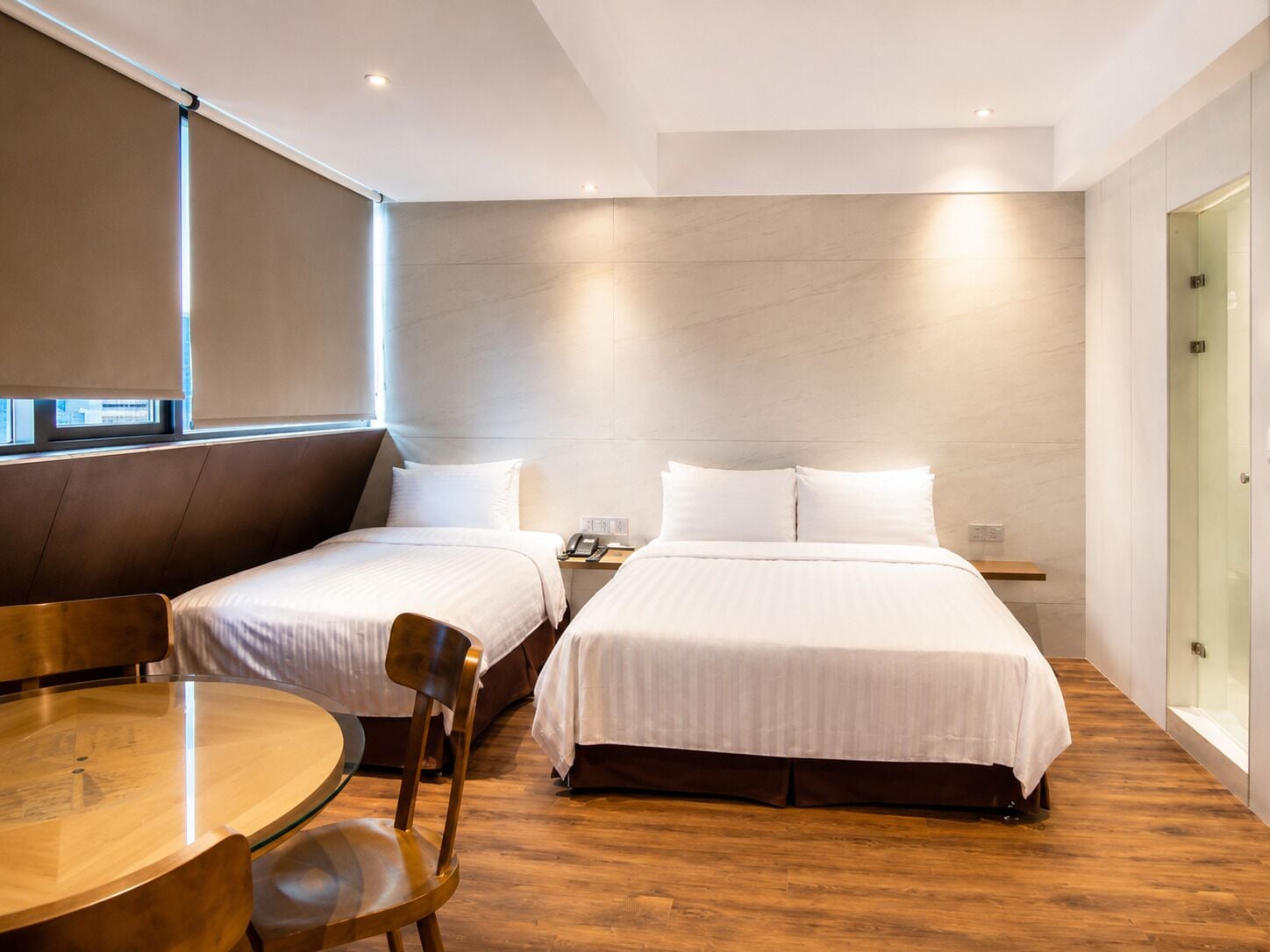 Una habitación de hotel moderna con dos camas, piso de madera, una mesa redonda y cabina de ducha de vidrio.