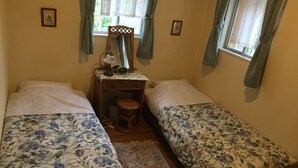 Et hyggeligt, lille værelse med to senge, et træskrivebord med stol, et lille bord med lampe, og et vindue med gardiner.
