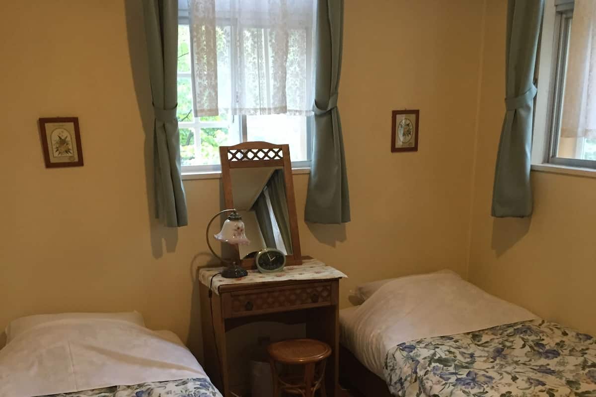 Ein Schlafzimmer mit zwei Einzelbetten, einem Holznachttisch mit Spiegel, einem kleinen Hocker und einem Fenster mit Vorhängen.