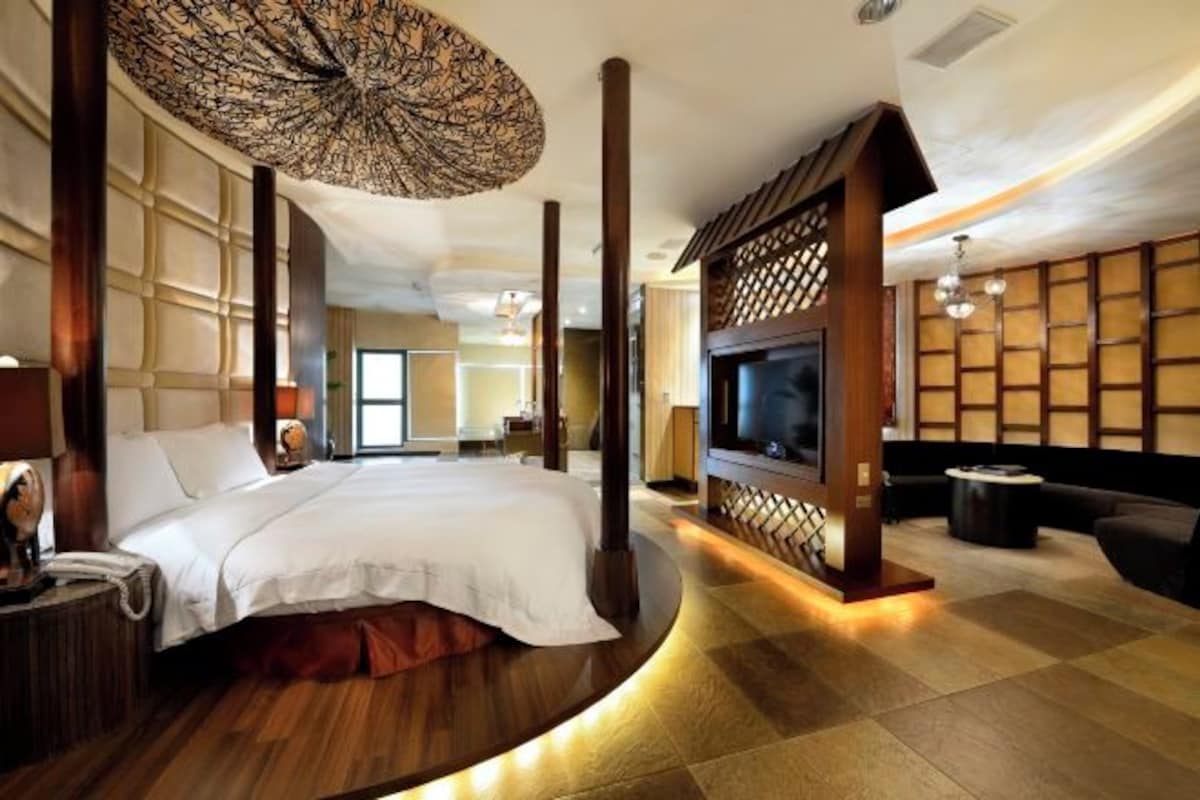 A modern hotel room with a curved bed, a television, a sofa, and a decorative ceiling light.