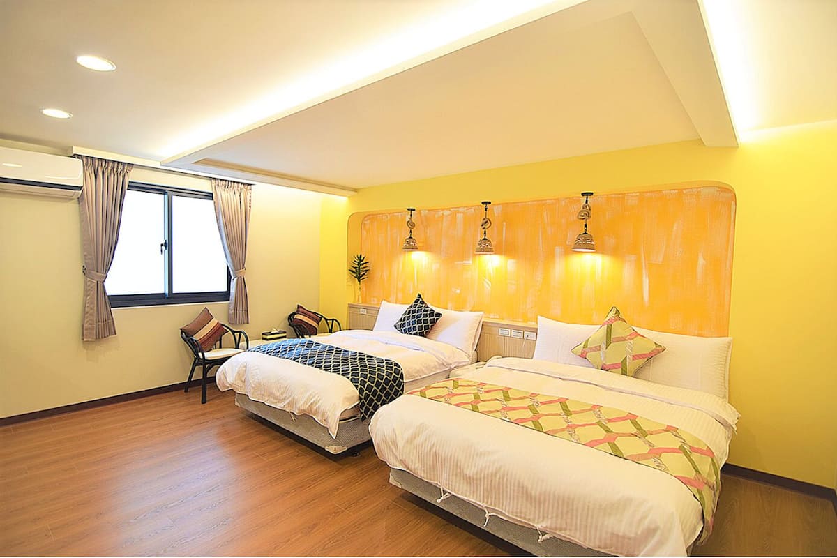A hotel room with two beds, wooden flooring, a window with curtains, and a yellow accent wall.