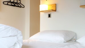 Uma cama bem arrumada com lençóis brancos, um luminária fixada na parede e uma pequena tomada elétrica.