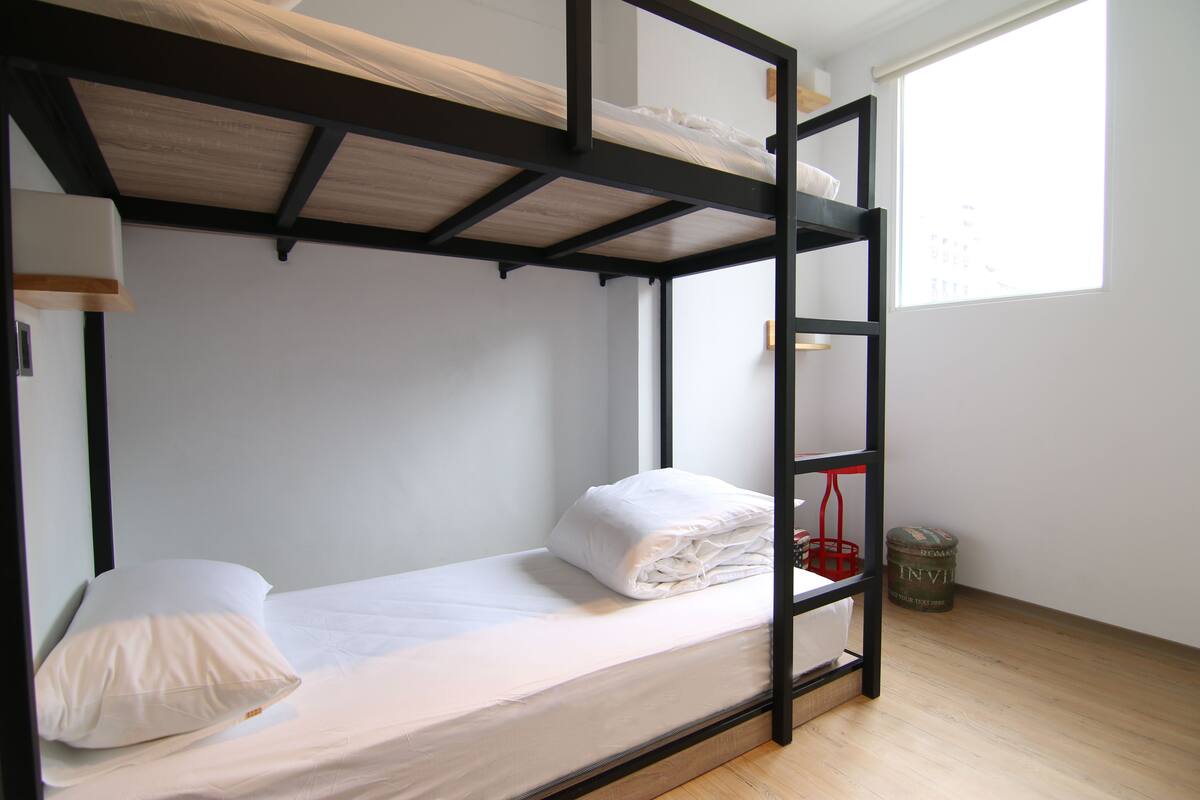 Ein Stockbett mit Holzrahmen und weißer Bettwäsche. Auf dem Boden befinden sich ein kleiner roter Korb und ein zylindrisches Gefäß.