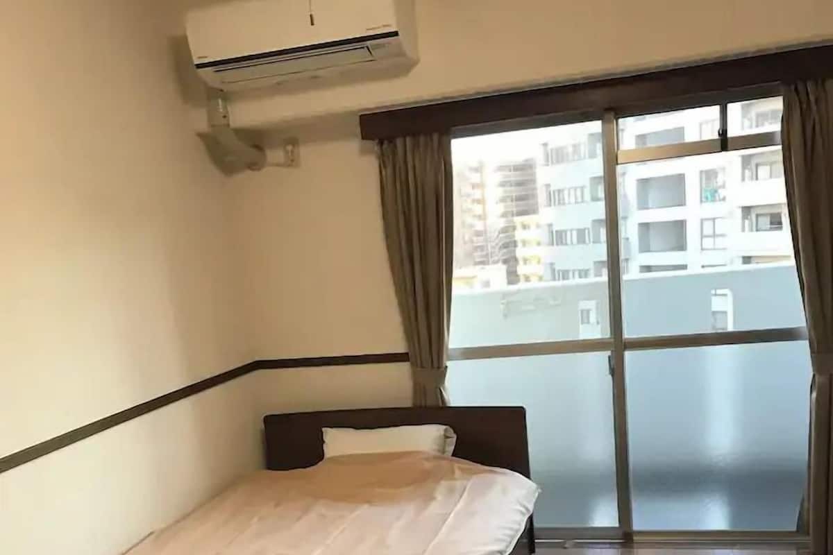 A hotel room with a bed, an air conditioning unit, a large window with curtains, and a view of buildings outside.