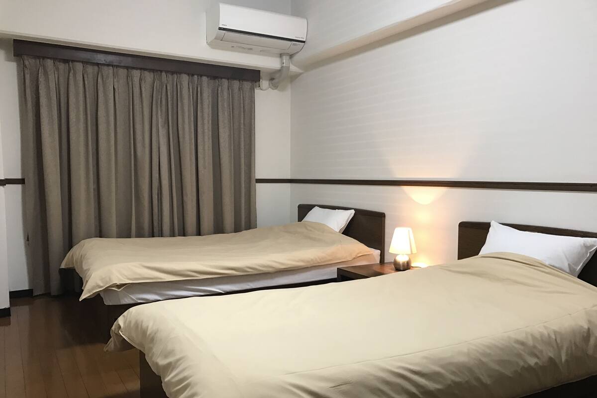 A hotel room with two beds, a nightstand with a lamp, a window with curtains, and an air conditioning unit.