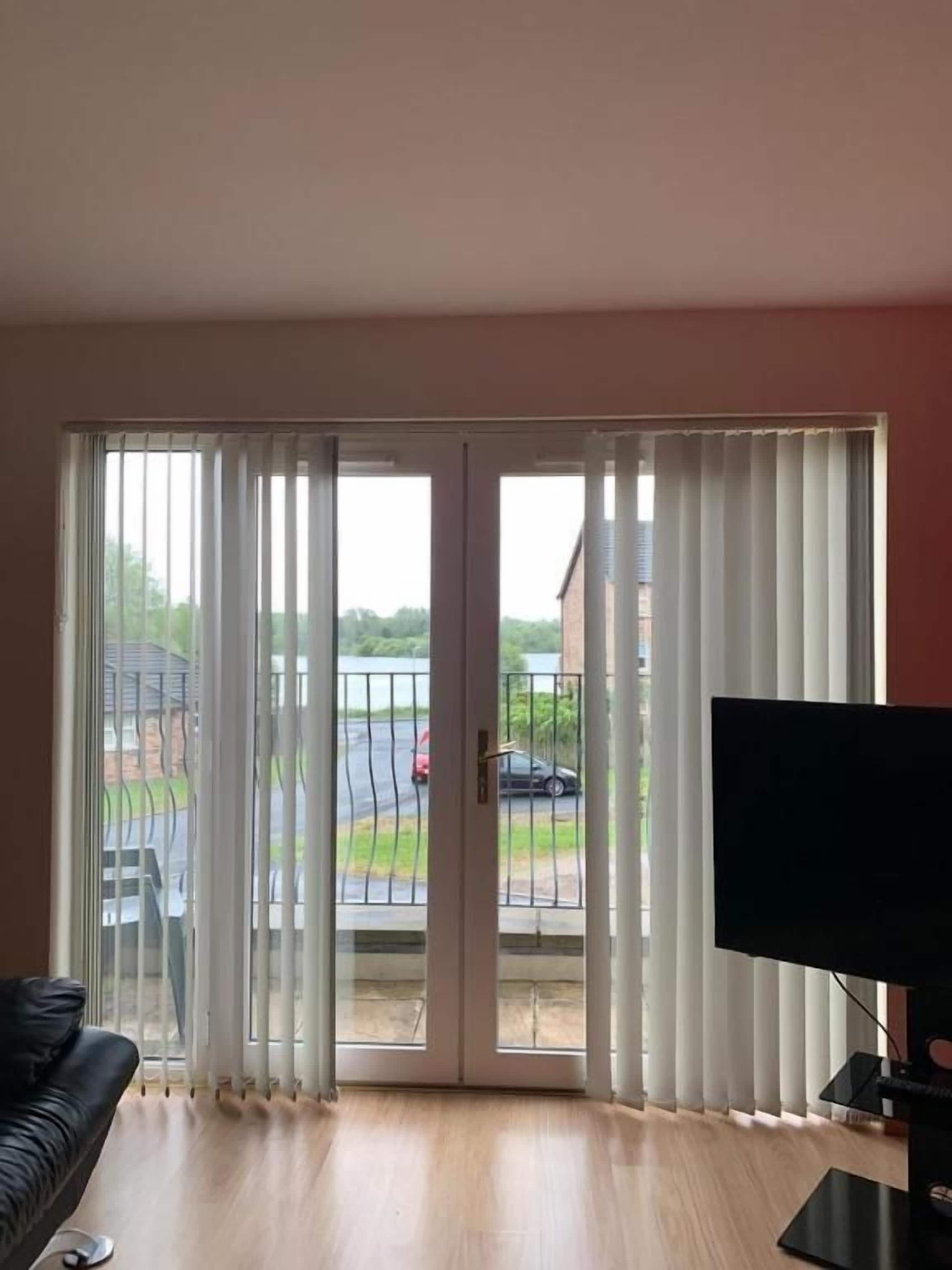 A room with a sliding glass door leading to a balcony, a flat-screen TV, and a wooden floor.