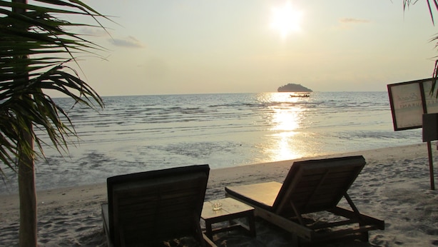 Une plage avec deux chaises longues en bois et une petite table, un palmier et un panneau indicateur en arrière-plan.