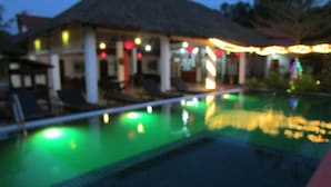 Une piscine dont l’eau reflète les lumières, un bâtiment avec un toit de chaume et quelques chaises longues.