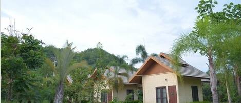 A bungalow-style building with a sloped roof and a beige exterior, surrounded by lush greenery and palm trees.