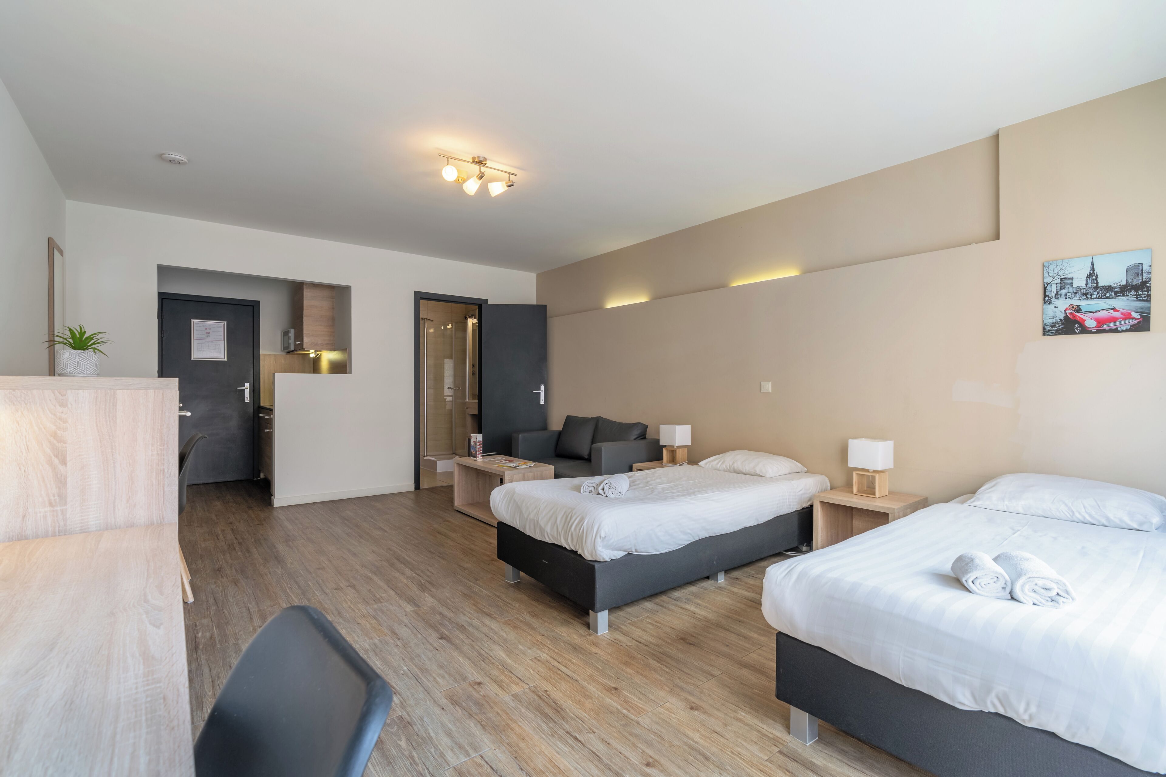 A modern hotel room with a wooden floor, a desk, a chair, two beds, a sofa, and a small table with a lamp.