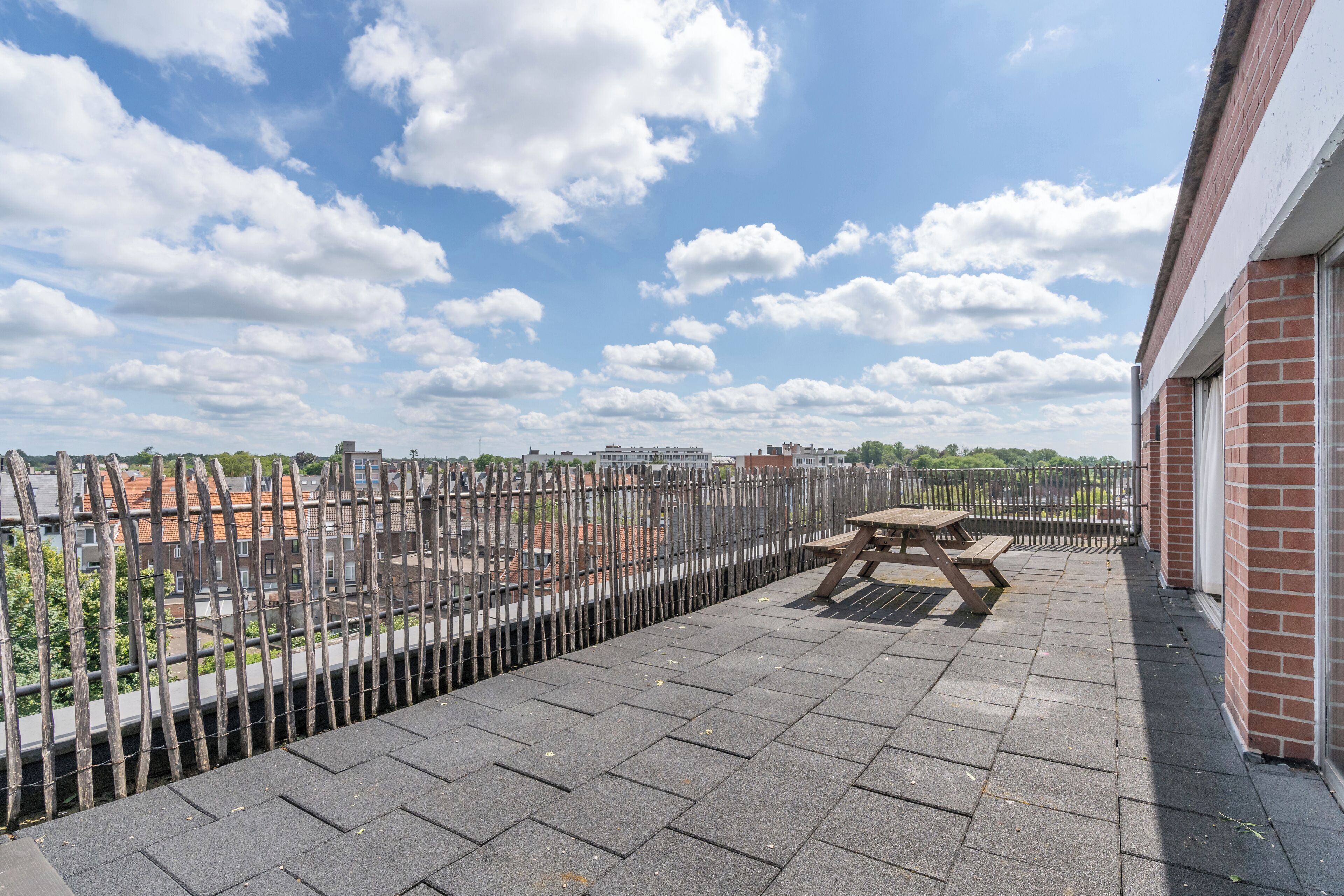 A rooftop terrace with a metal fence, a wooden table, and a brick building in the background.