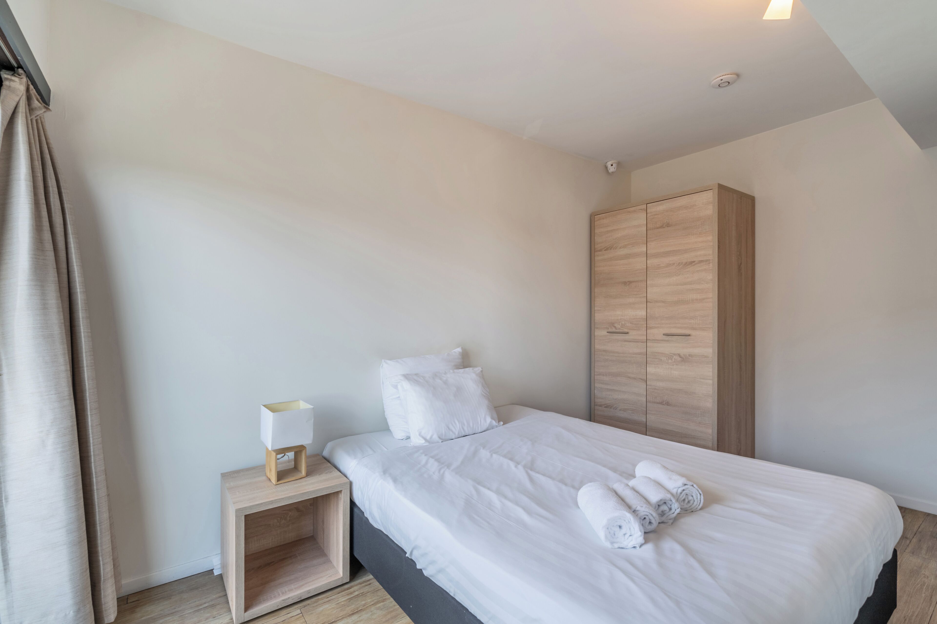Um quarto moderno com uma cama grande, um guarda-roupa de madeira, uma mesa de cabeceira com abajur e toalhas dobradas com cuidado.
