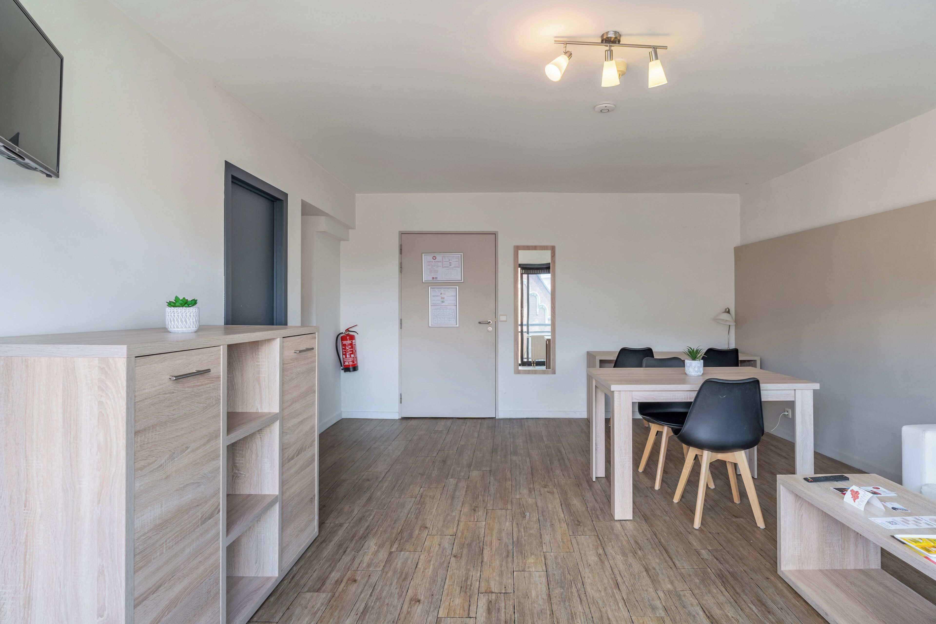 A modern, well-lit room with a wooden floor, a desk with chairs, a small table with a plant, and a fire extinguisher mounted on the wall.