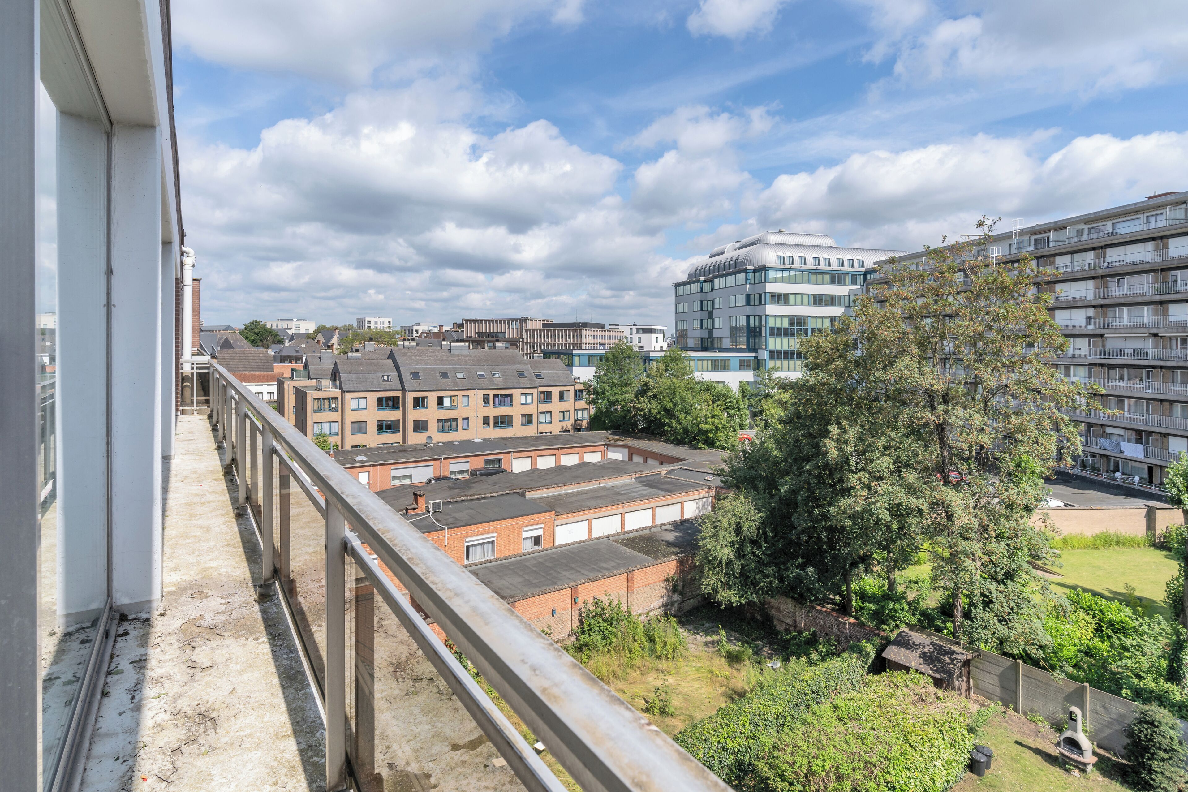 A view from a balcony overlooking a residential area with modern and older buildings, a grassy park, and a clear sky.