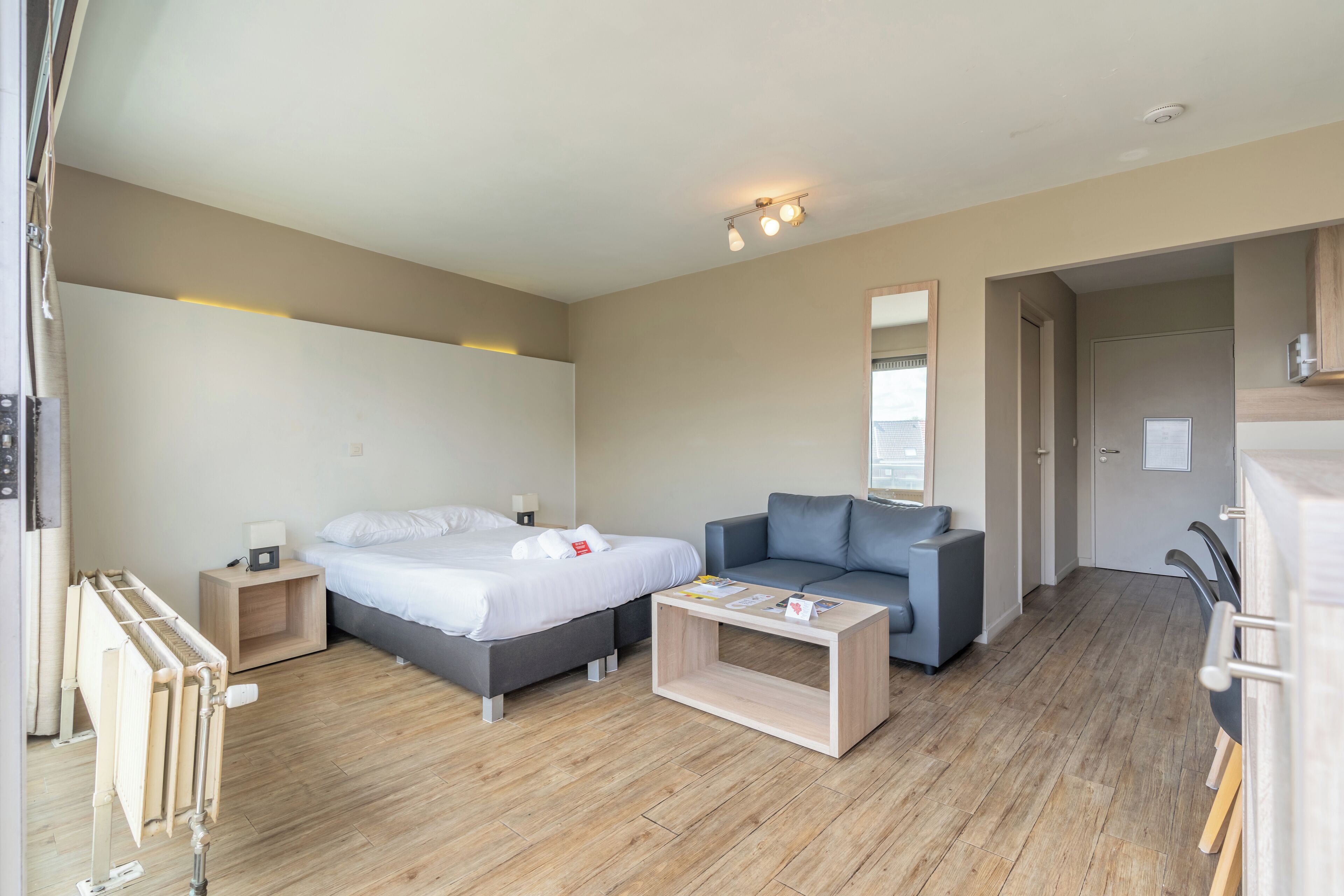 Una habitación de hotel moderna con cama, sofá, una mesita de centro de madera y una mesita de noche pequeña con lámpara.