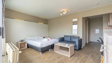 A modern hotel room with a bed, a sofa, a wooden coffee table, and a small bedside table with a lamp.