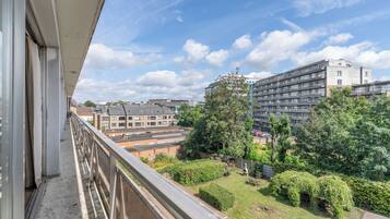A balcony with a view of a residential area, including a multi-story building and a park with trees and a bench.