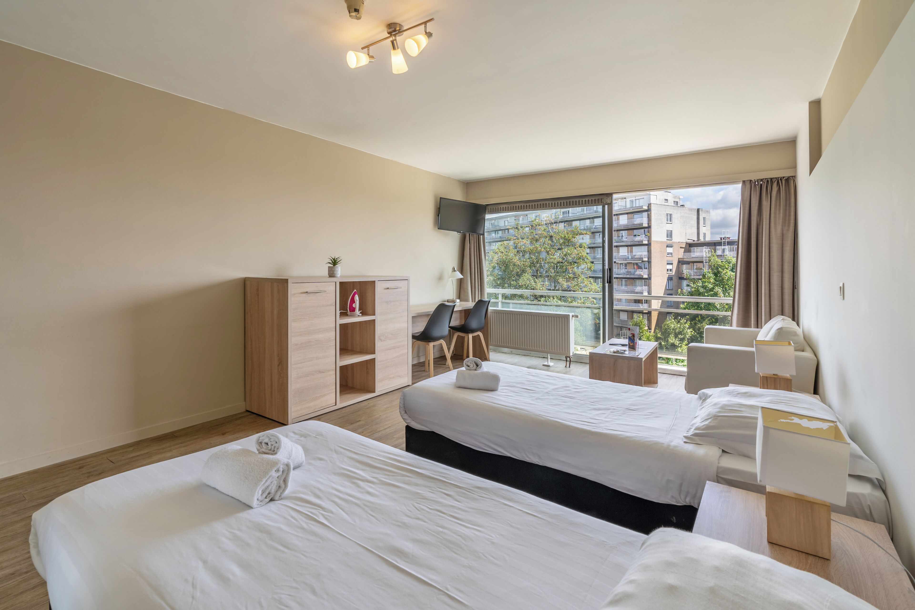 A hotel room with two beds, a desk, a chair, a TV, and a window with a view of buildings.