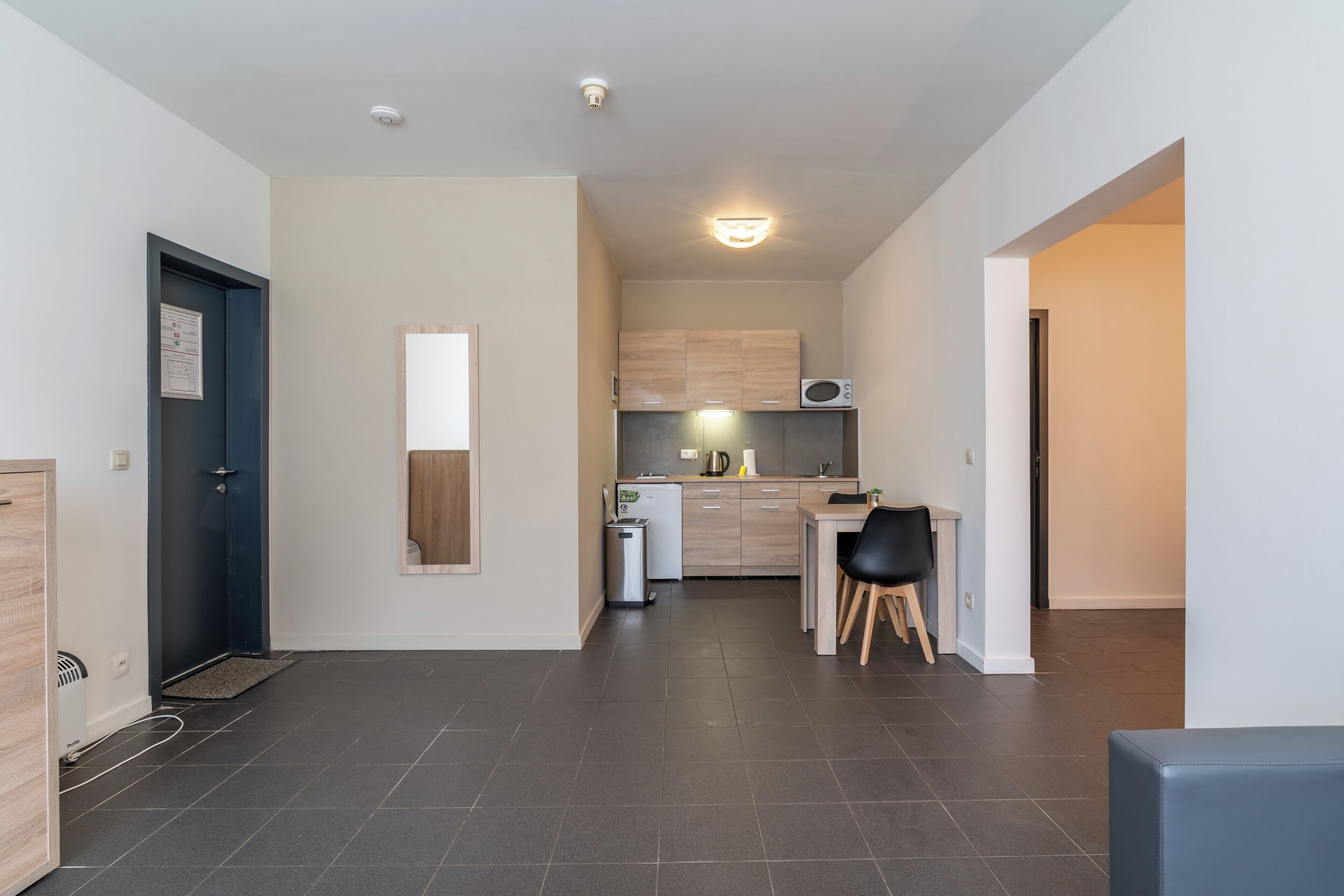 Una cocina moderna con baldosas oscuras, una zona de comedor con una silla negra y un refrigerador pequeño.