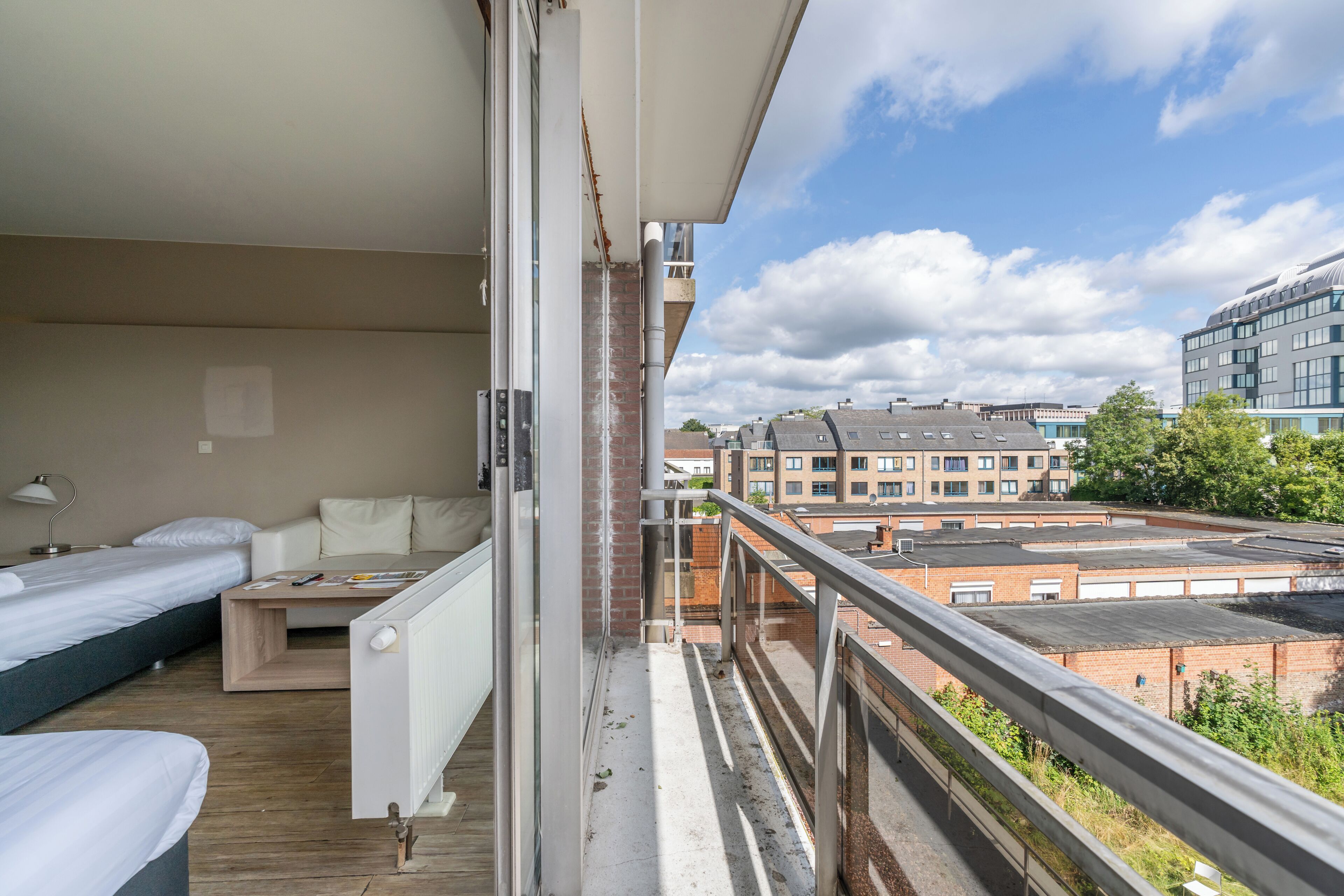 A balcony with a view of the cityscape, a bed, a small table, and a radiator.