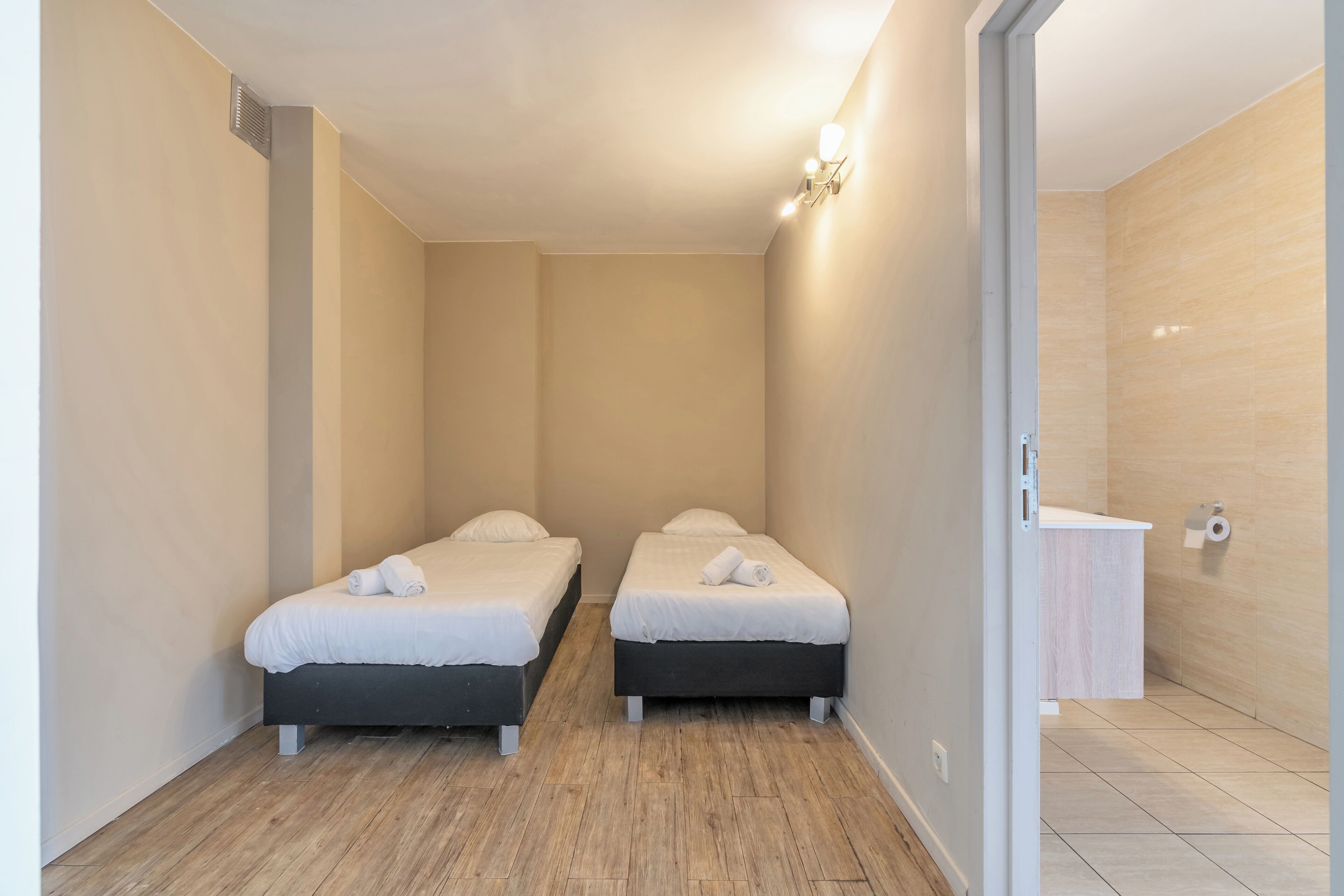 Una habitación pequeña y limpia con dos camas, piso de madera, pared de color claro y un baño pequeño con lavamanos e inodoro.