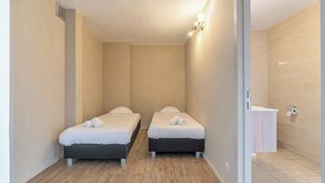 Une petite pièce propre, avec deux lits, un plancher en bois, un mur de couleur claire et une petite salle de bain comprenant un lavabo et des toilettes.