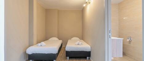 Una habitación pequeña y limpia con dos camas, piso de madera, pared de color claro y un baño pequeño con lavamanos e inodoro.