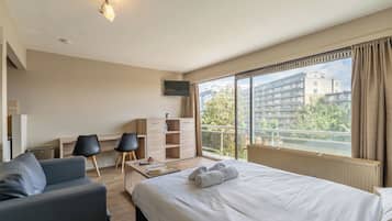 Habitació d'hotel amb un llit gran, un escriptori, dues cadires, un televisor i un balcó amb vista a edificis i arbres.