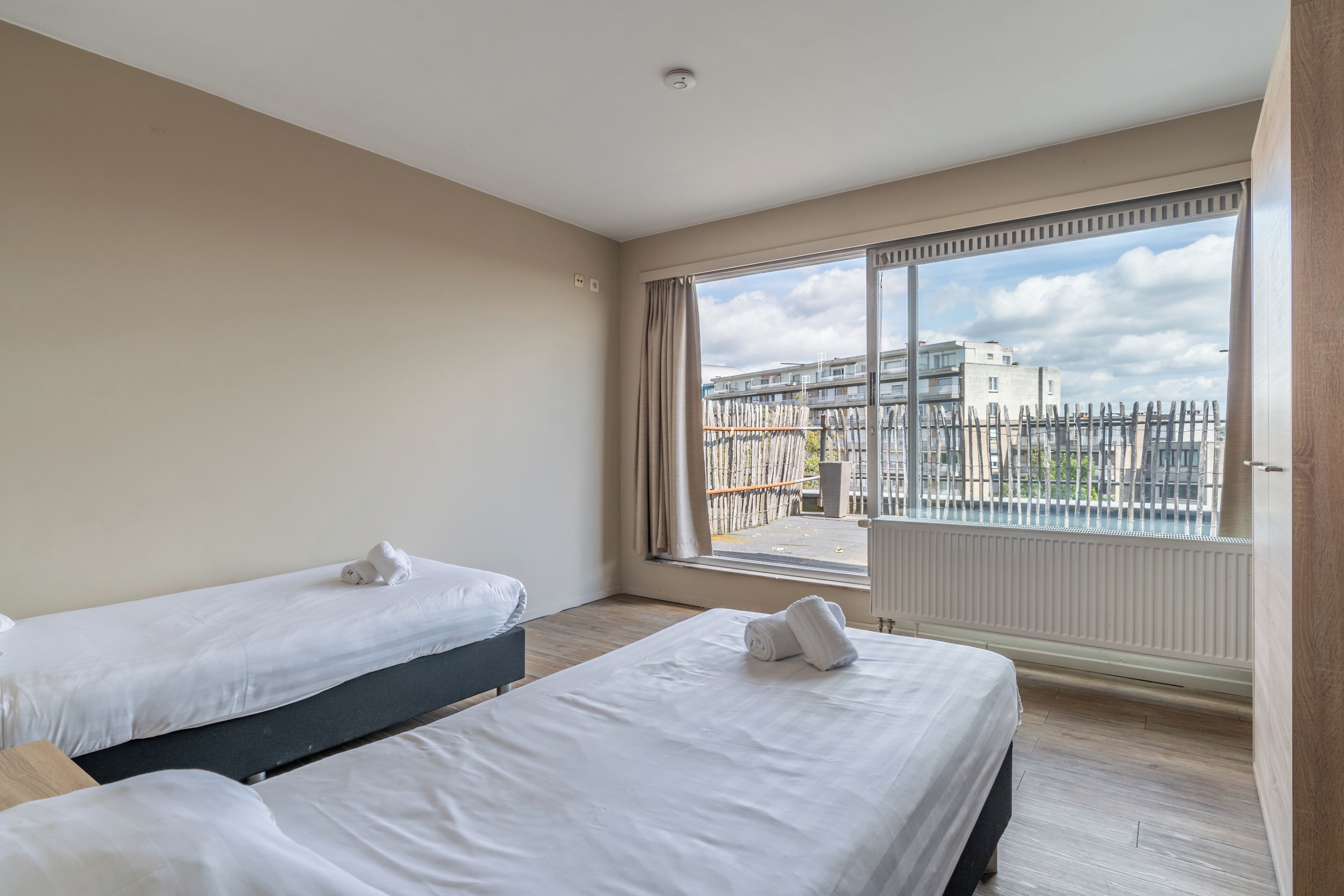 Une chambre d’hôtel avec deux lits, une grande fenêtre donnant sur des immeubles et une clôture, et un radiateur fixé au mur.