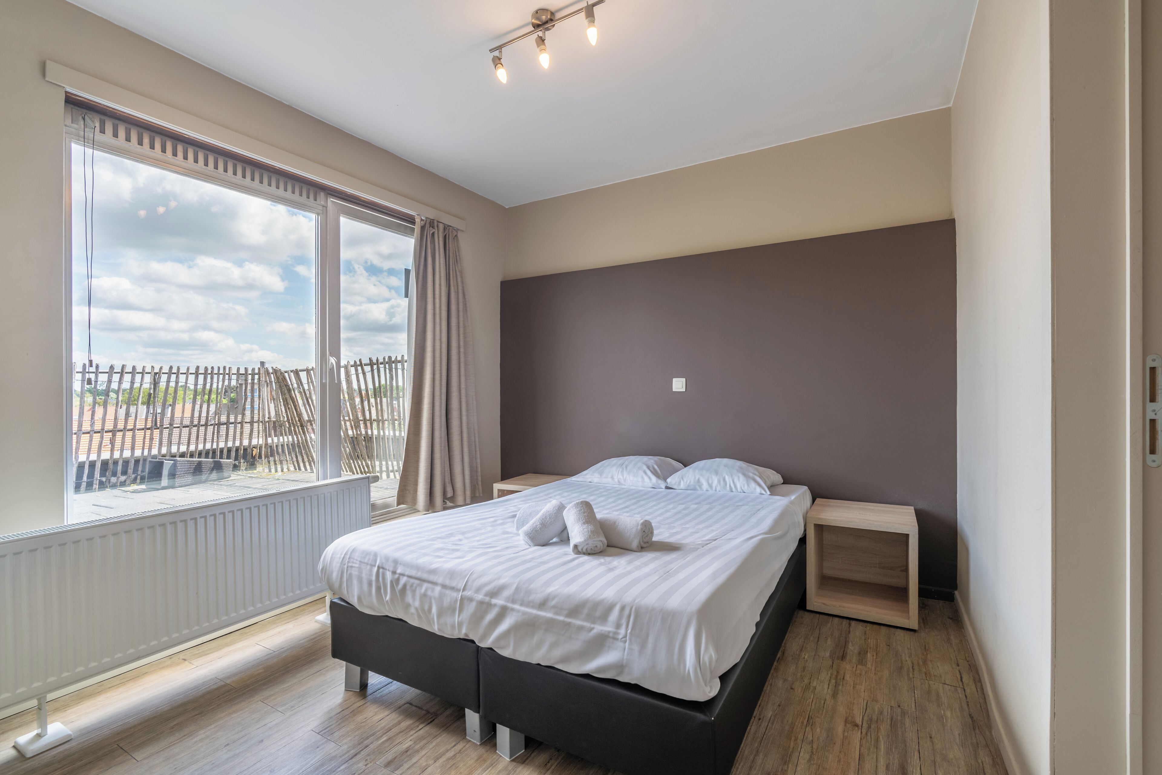 A bedroom with a large window, a bed with white linens, a dark headboard, and a small bedside table.