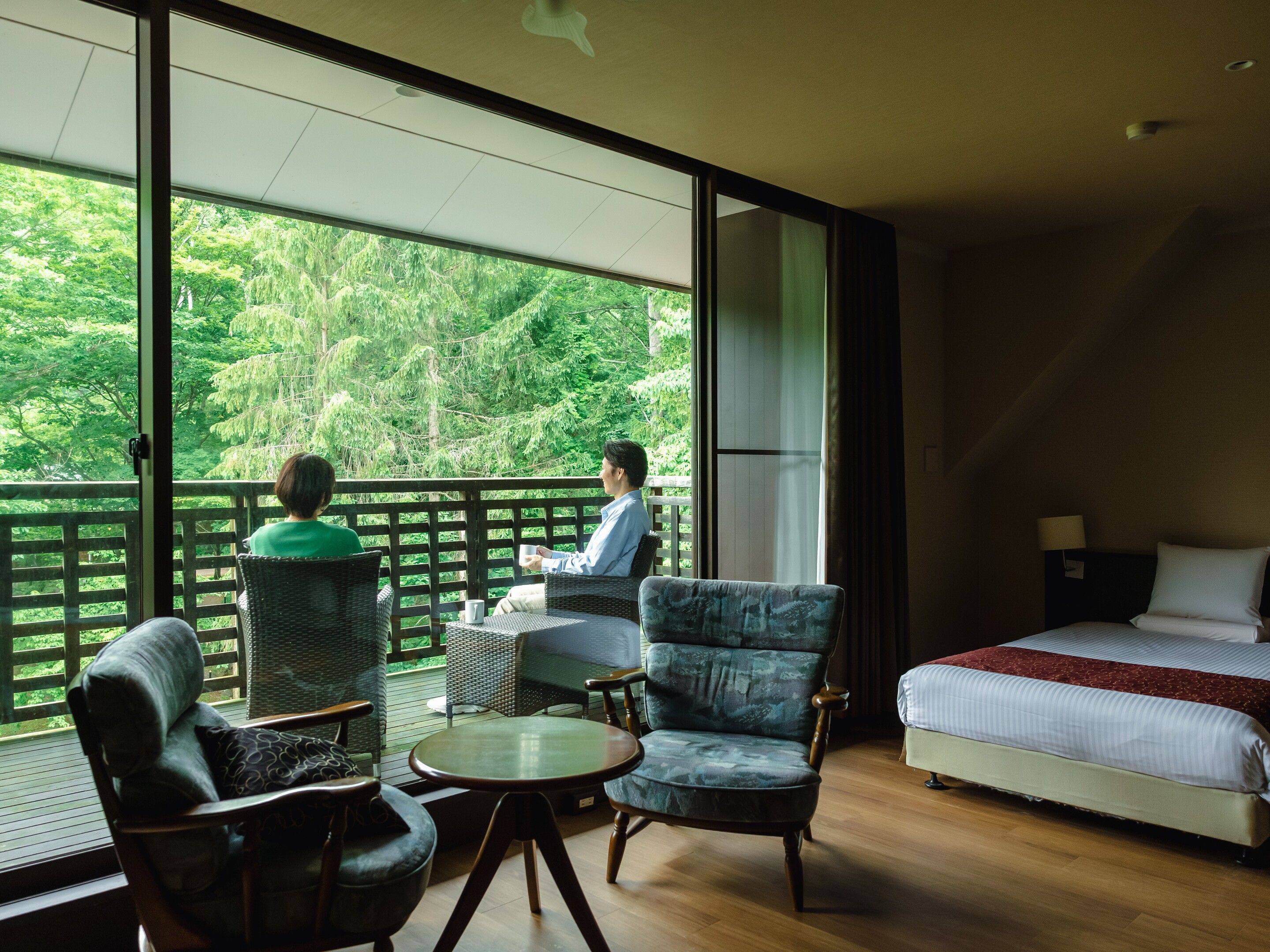 A room with a bed, a table, and two chairs. There is a balcony with a view of trees.
