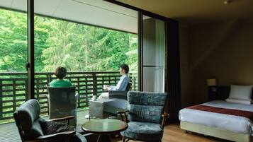 A room with a bed, a table, and two chairs. There is a balcony with a view of trees.