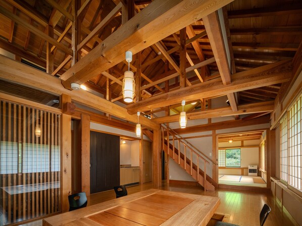 A spacious room with a high ceiling, wooden beams, a dining table, and a staircase leading to another level.