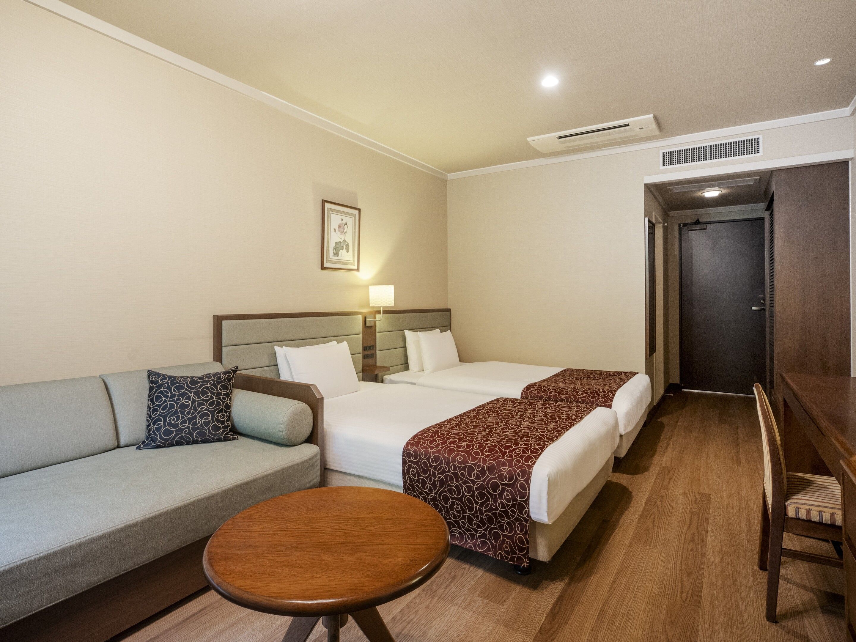 A hotel room with a sofa, two beds, a small table, and a door.