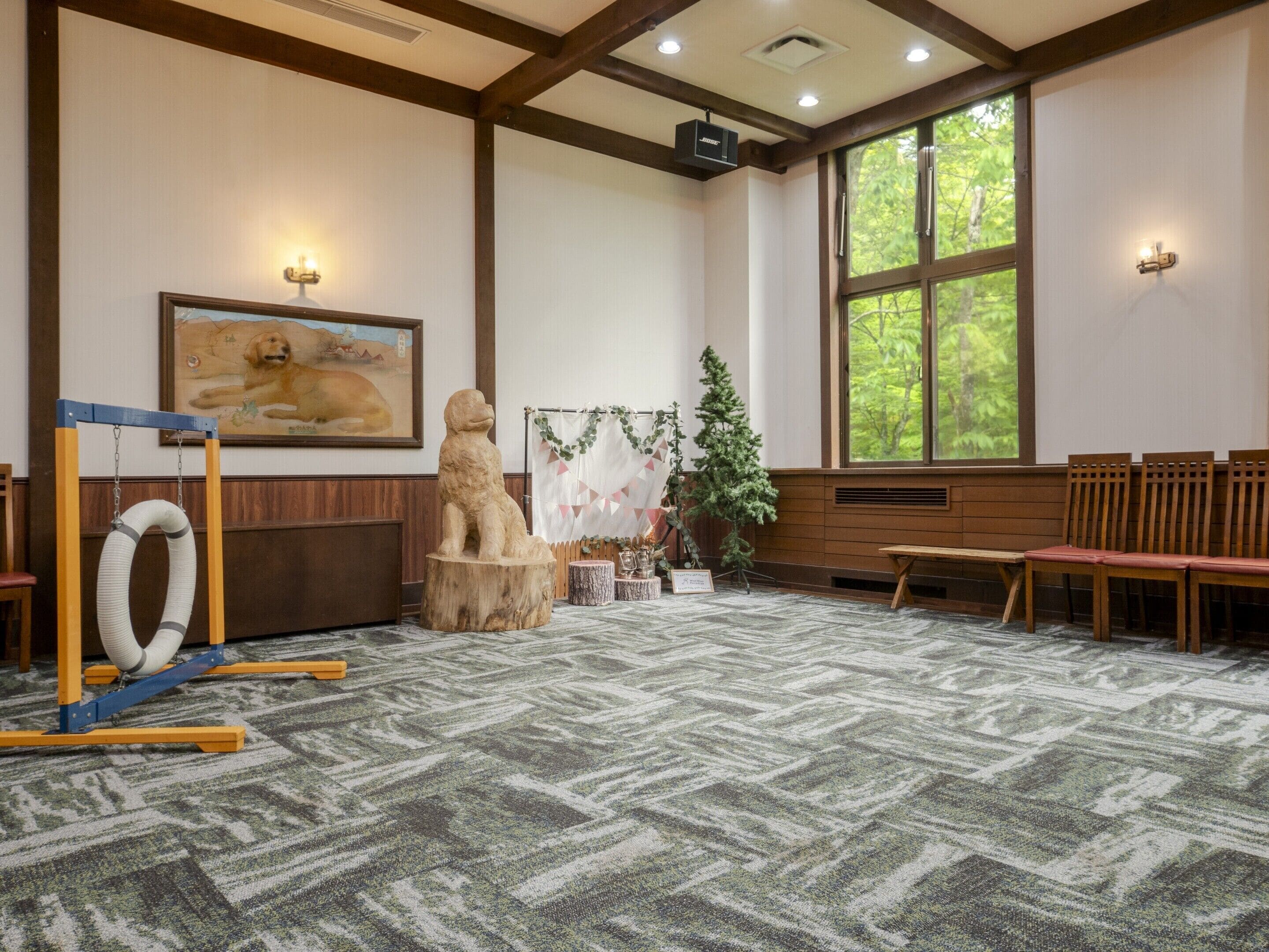 A room with a patterned carpet, wooden benches, a framed painting, and a window with a view of trees.