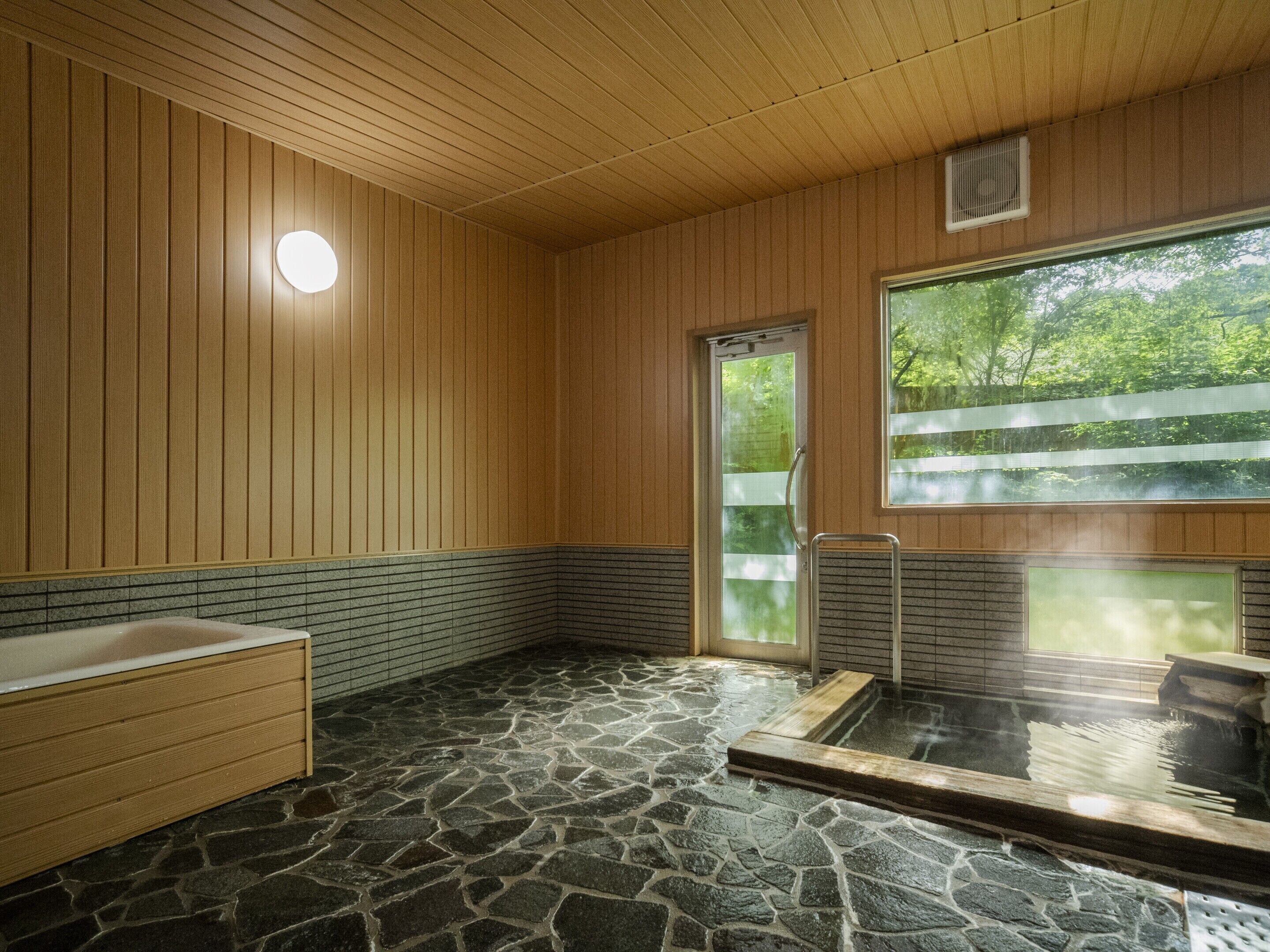 A traditional Japanese-style bath with a stone floor, wooden walls, and a large window.